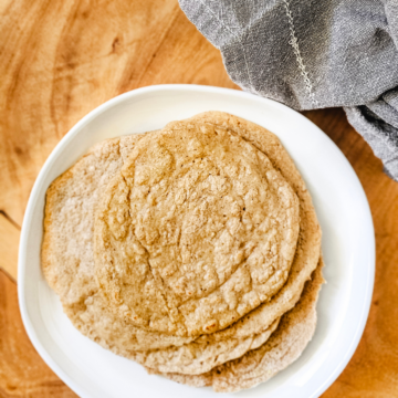 Vertical overhead image of a stack of gluten-free vegan oat tortillas on a white plate on a wooden board next to a grey linen napkin.