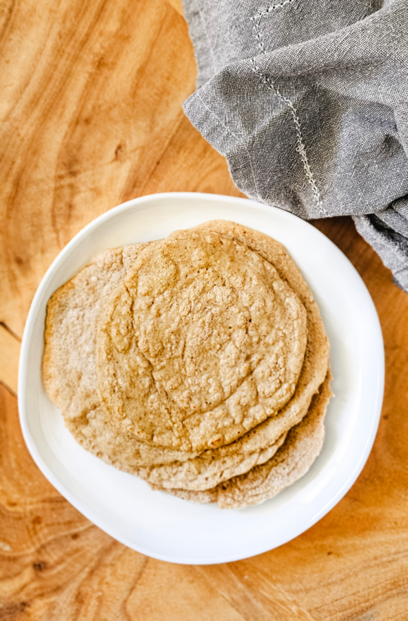 Vertical overhead image of a stack of gluten-free vegan oat tortillas on a white plate on a wooden board next to a grey linen napkin.