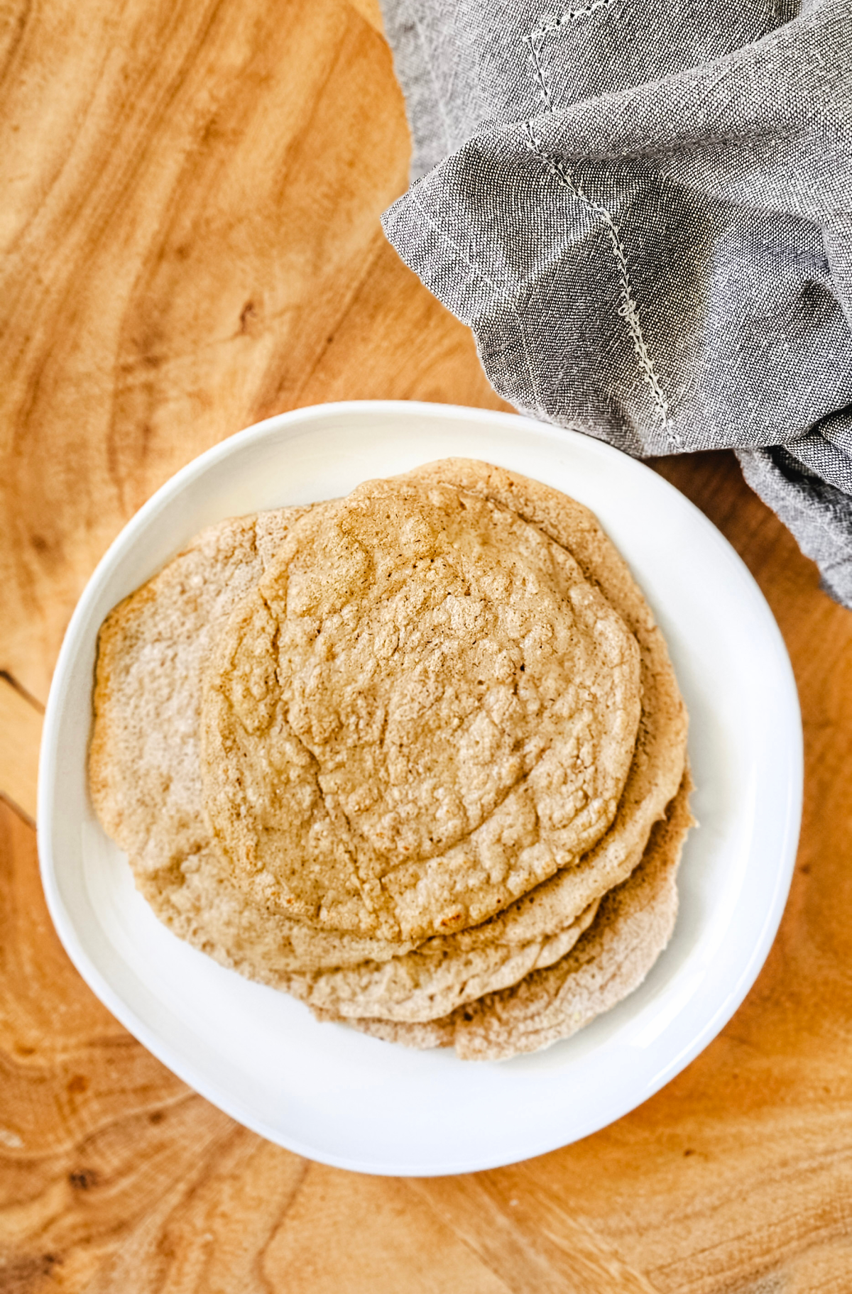 Vertical overhead image of a stack of gluten-free vegan oat tortillas on a white plate on a wooden board next to a grey linen napkin.