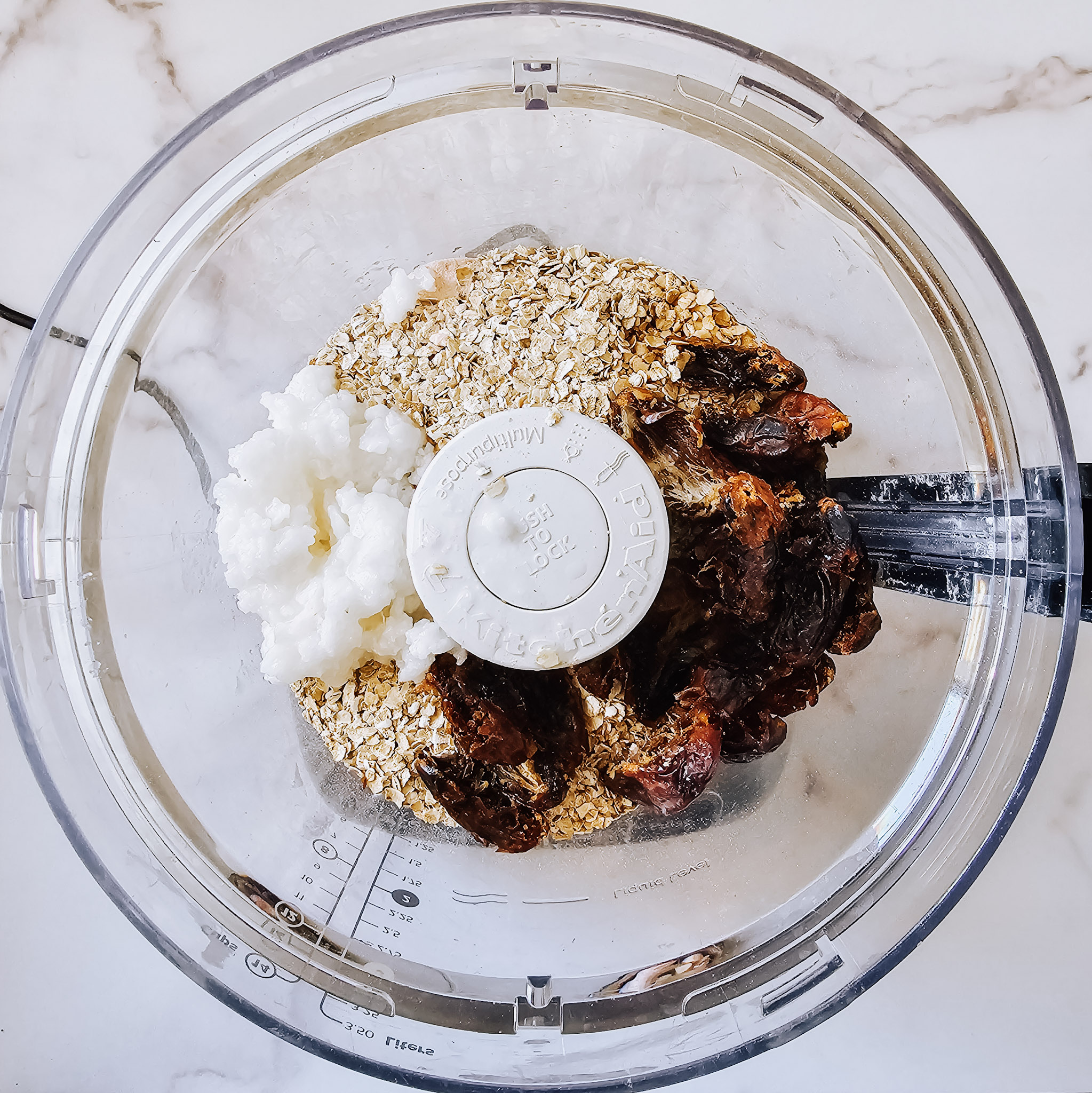 Overhead image of the ingredients needed to make gluten-free vegan no-bake chocolate peanut butter oat bars in a food processor on a white marble surface.