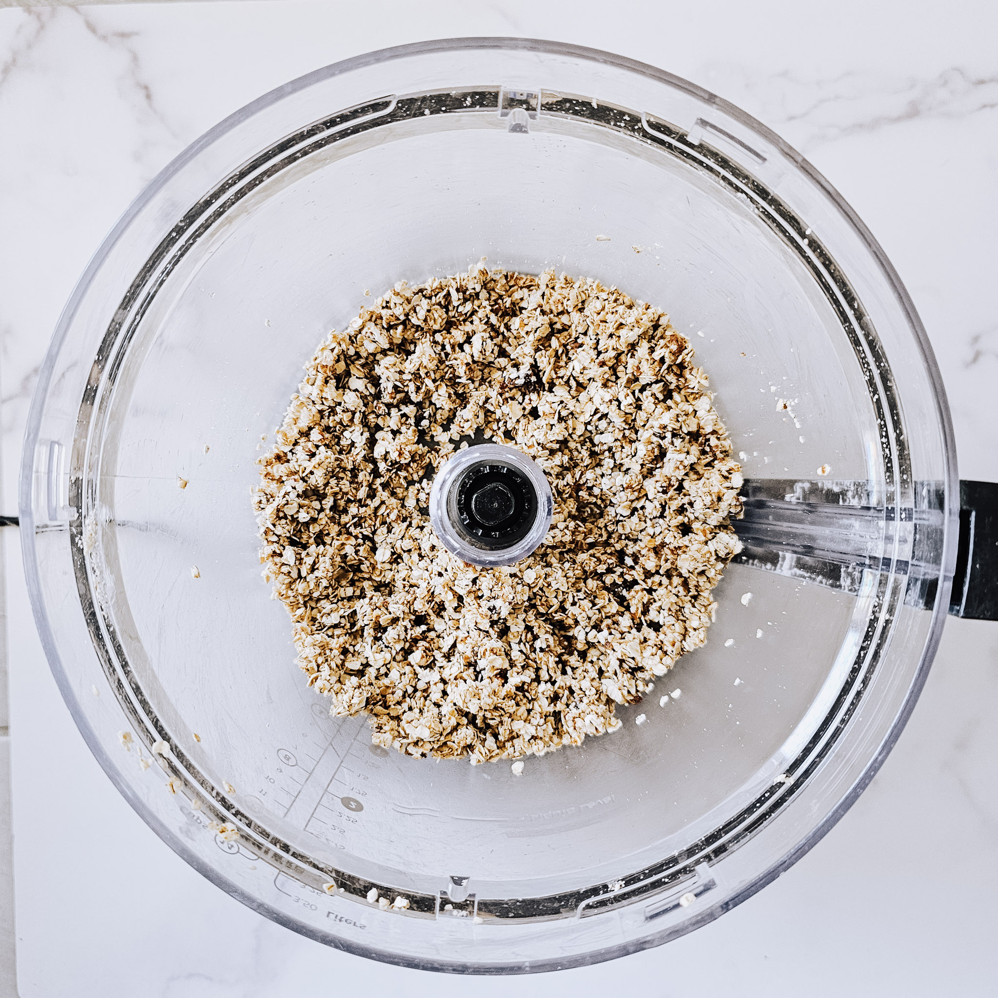 Overhead image of the ingredients needed to make the top and bottom layer of gluten-free vegan no-bake chocolate peanut butter oat bars blended in a food processor on a white marble surface.