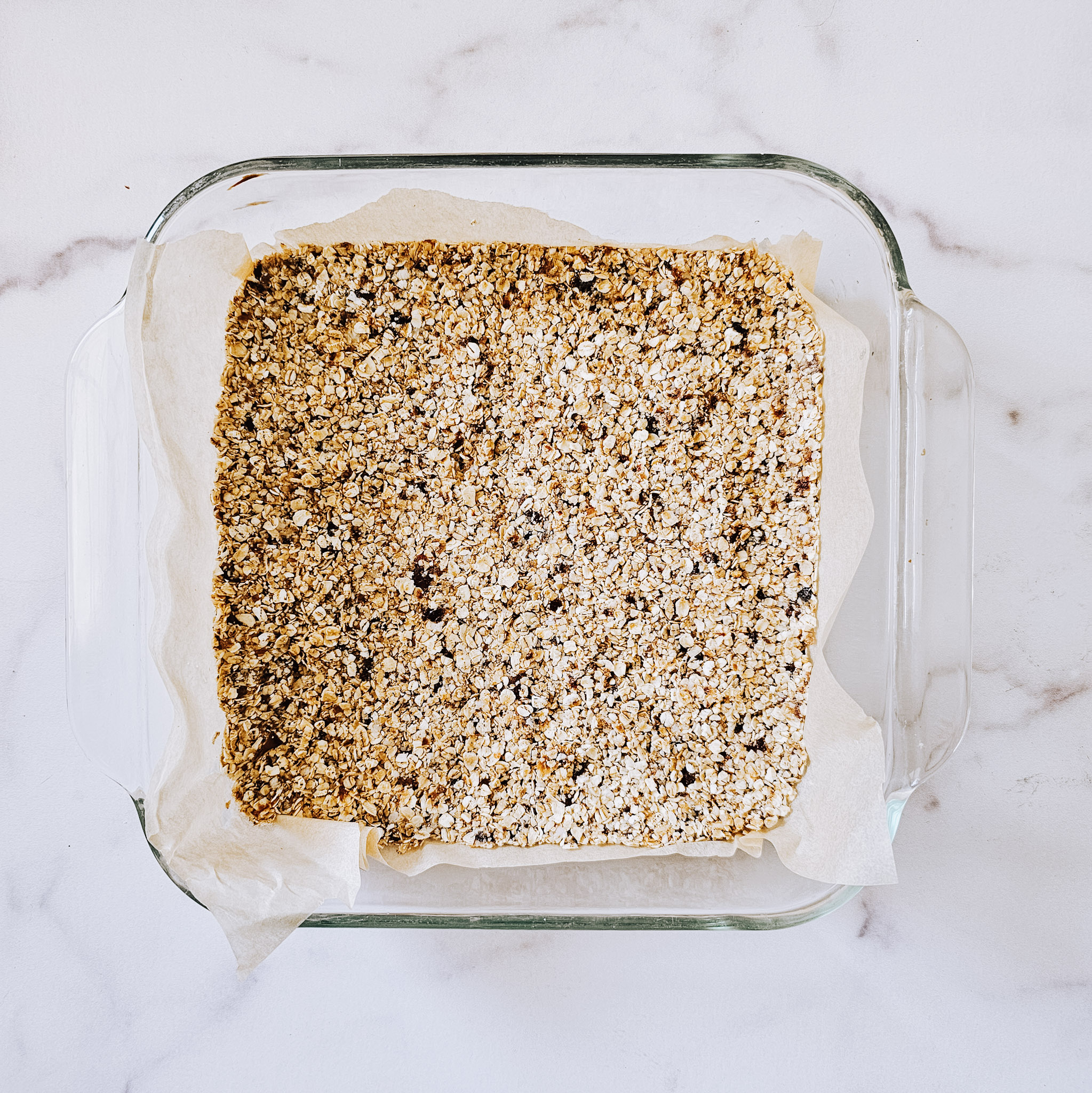 Overhead image of the ingredients for the bottom layer of gluten-free vegan no-bake chocolate peanut butter oat bars in a glass baking dish lined with parchment paper on a white marble surface.