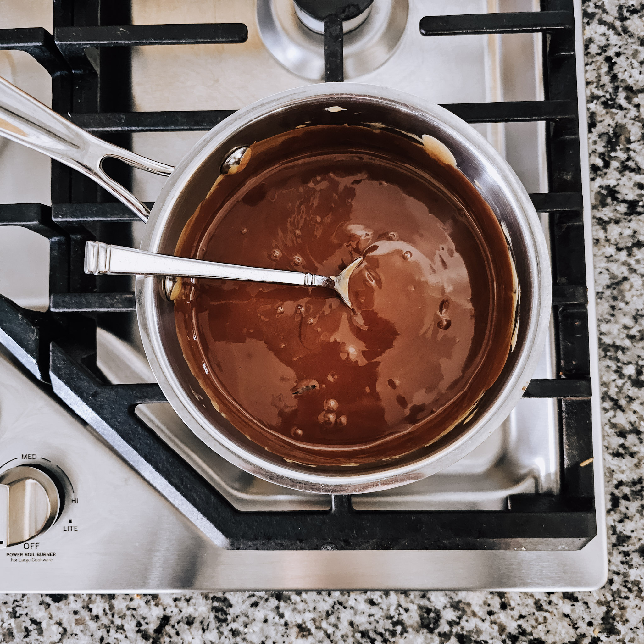 Overhead image of melted chocolate in a stainless steel saucepan on a gas cooktop.