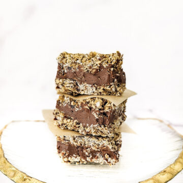 A stack of three gluten-free vegan no-bake chocolate peanut butter oat bars on a decorative gold rimmed glass plate on a white marble background.