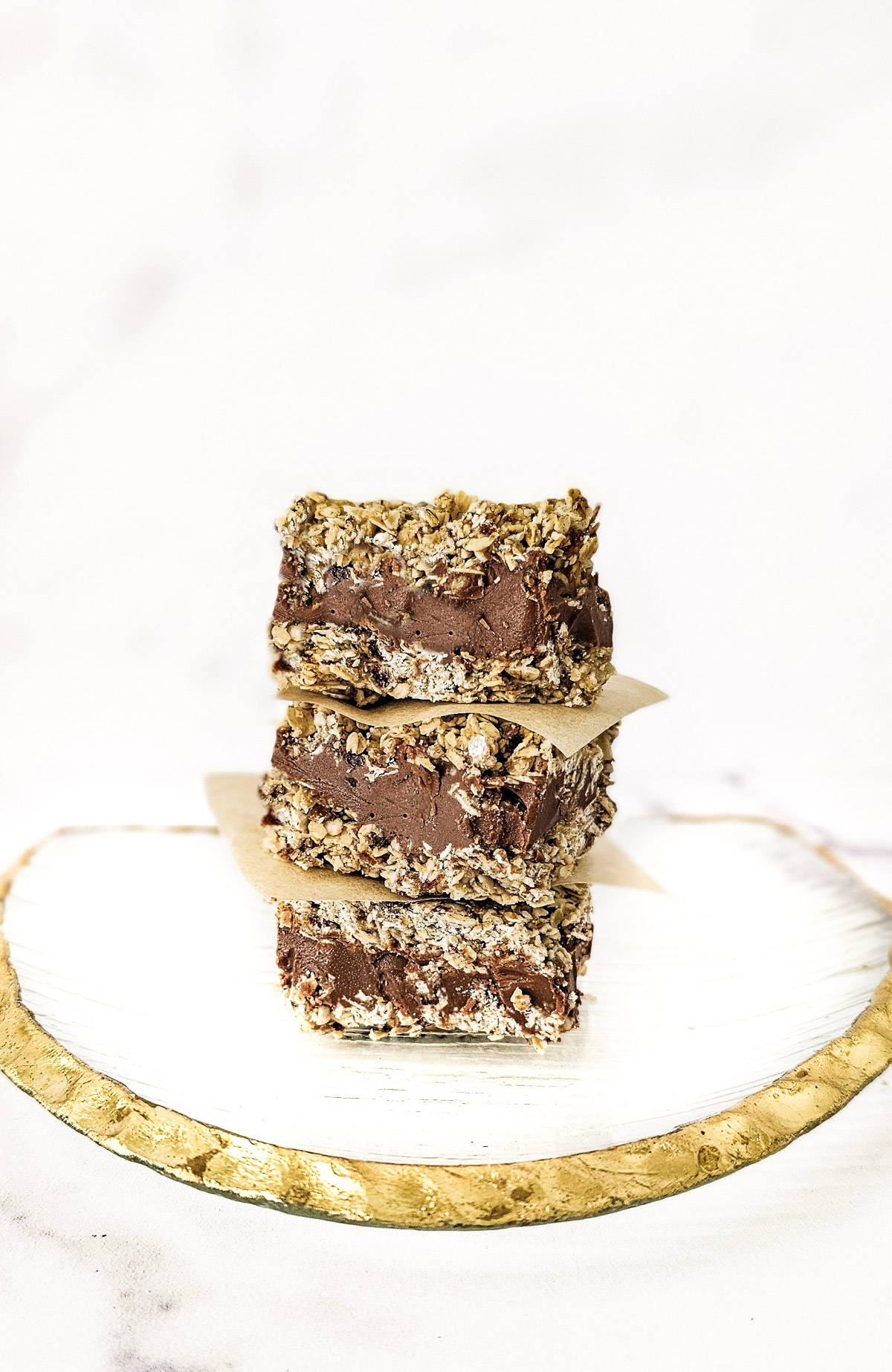 A stack of three gluten-free vegan no-bake chocolate peanut butter oat bars on a decorative gold rimmed glass plate on a white marble background.
