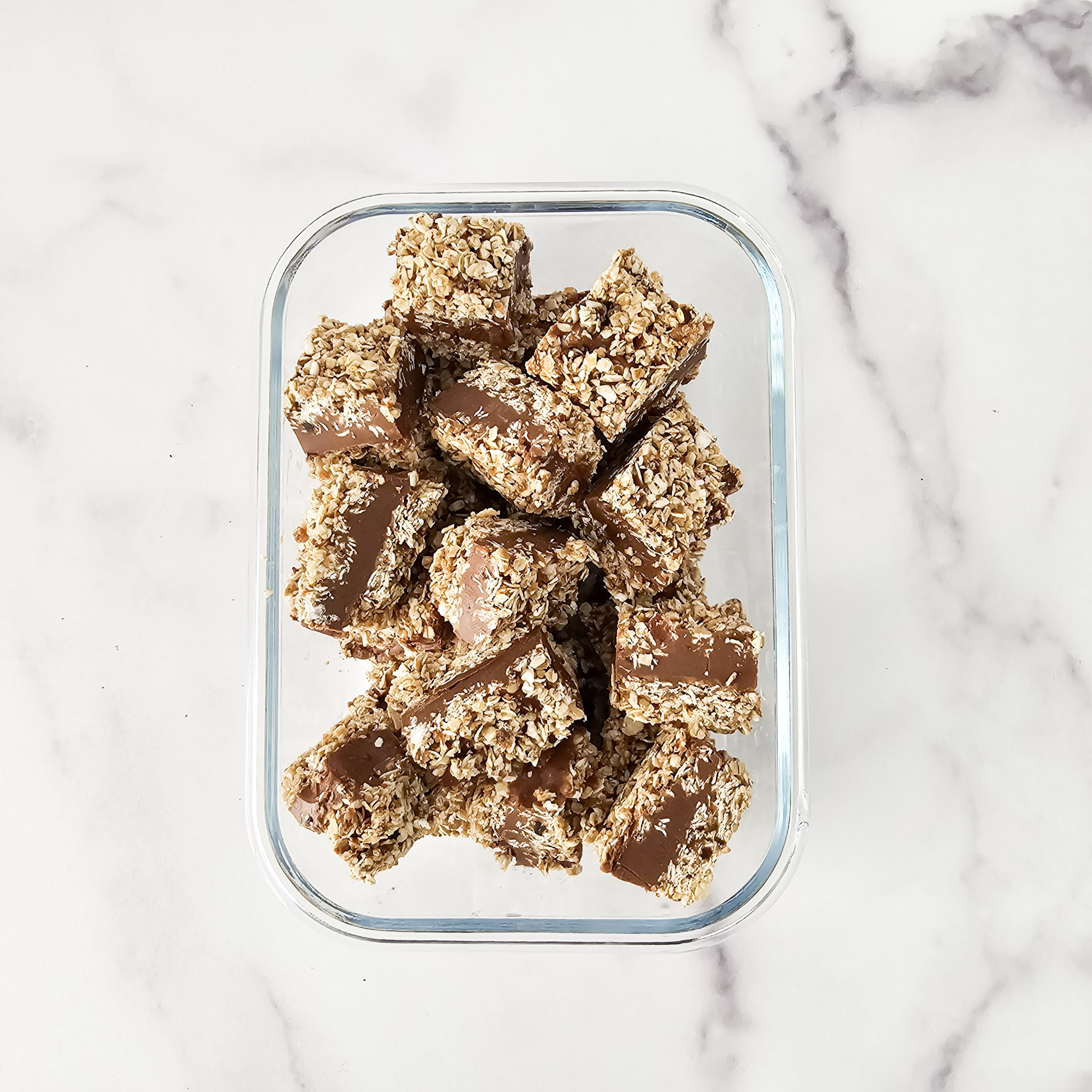 Overhead image of gluten-free vegan no-bake chocolate peanut butter oat bars cut into bite size pieces in a glass storage container on a white marble surface.