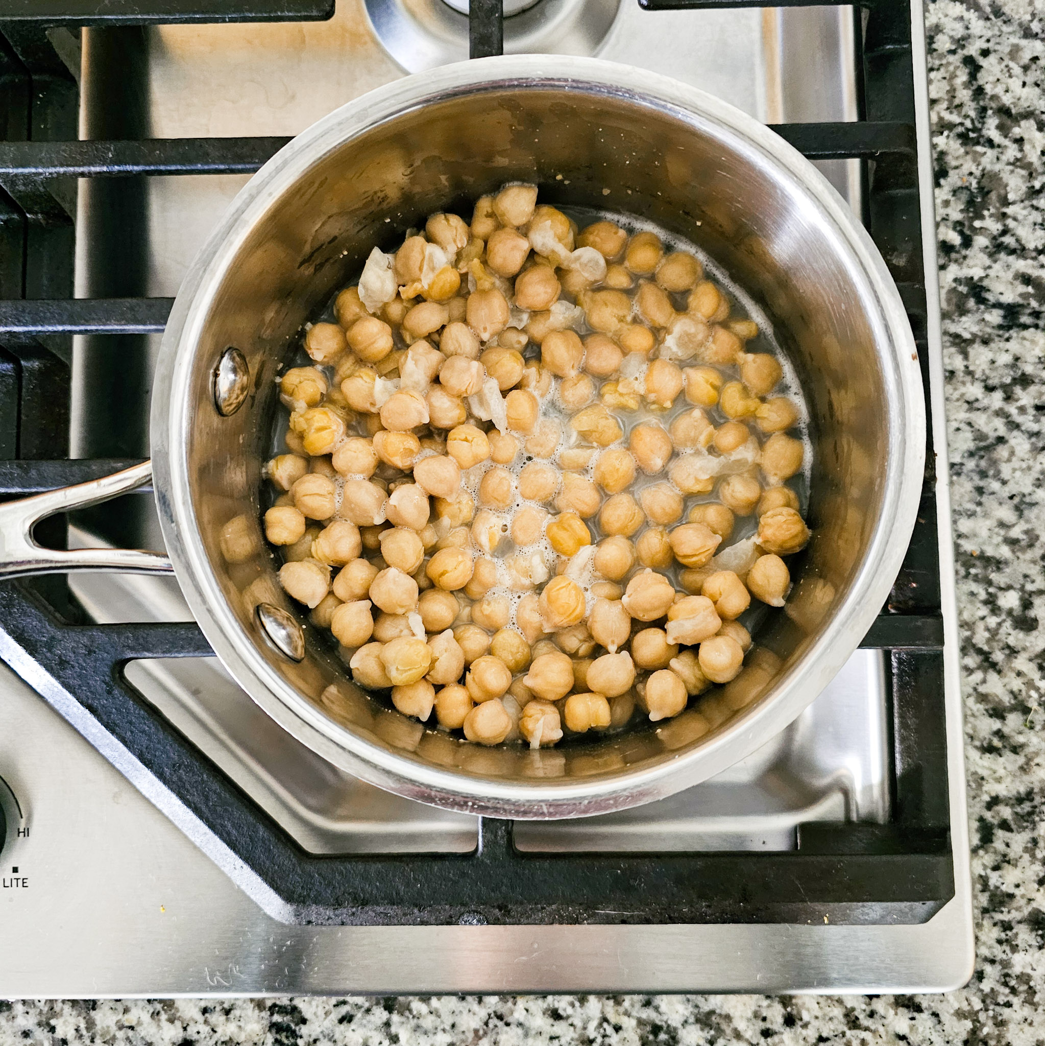 Ingredients to make the chickpea mixture of gluten-free vegan creamy musabaha being added to a stainless-steel saucepan on a gas cooktop.
