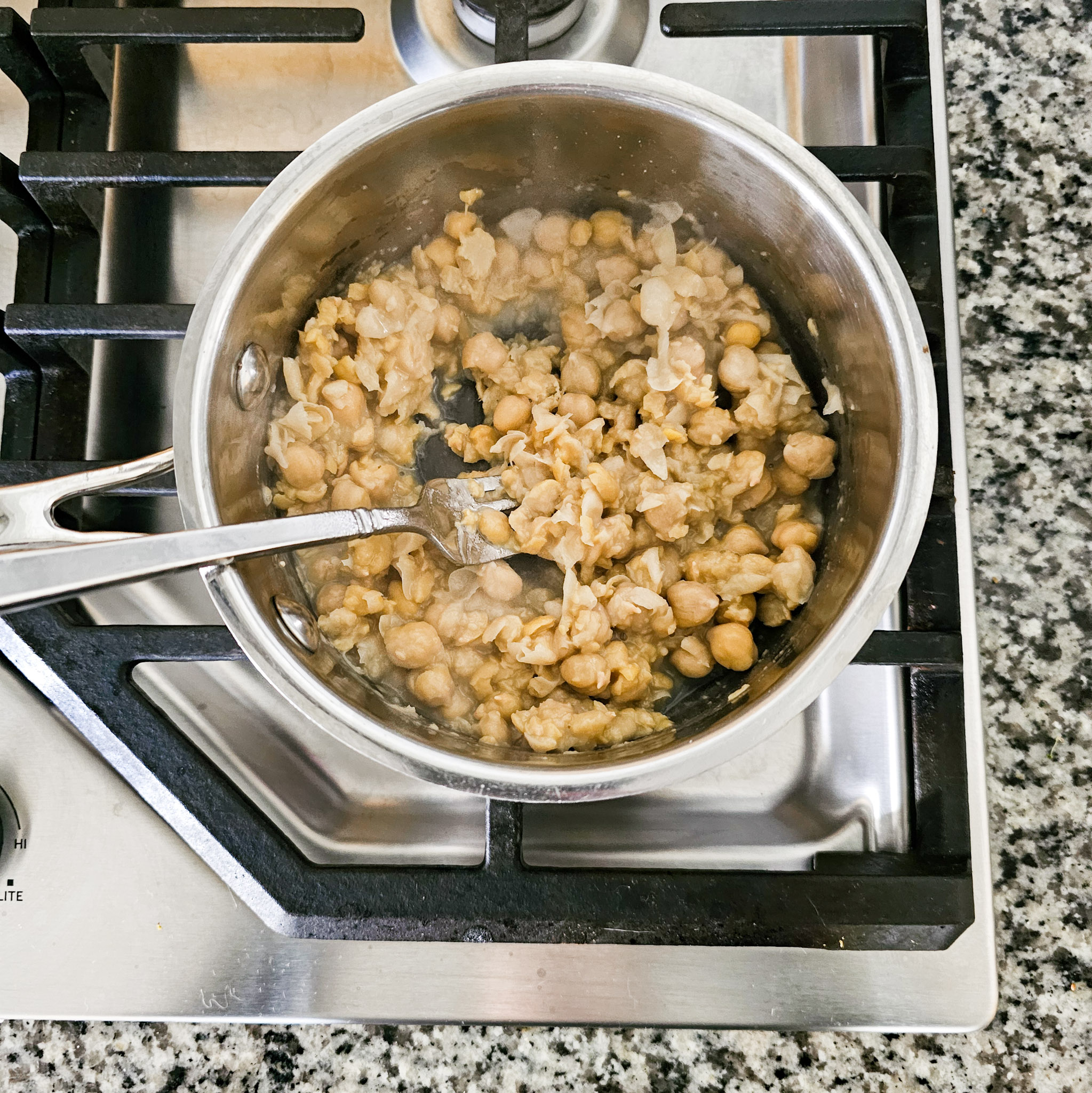 Ingredients to make the chickpea mixture of gluten-free vegan creamy musabaha being mashed together with a fork in a stainless-steel saucepan on a gas cooktop.