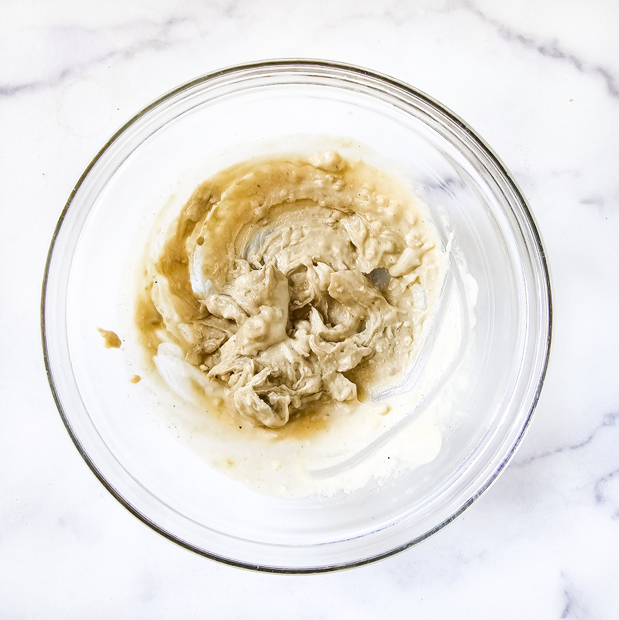 Overhead image of the ingredients needed to make gluten-free vegan creamy musabaha mixed together in a glass bowl on a white marble surface.