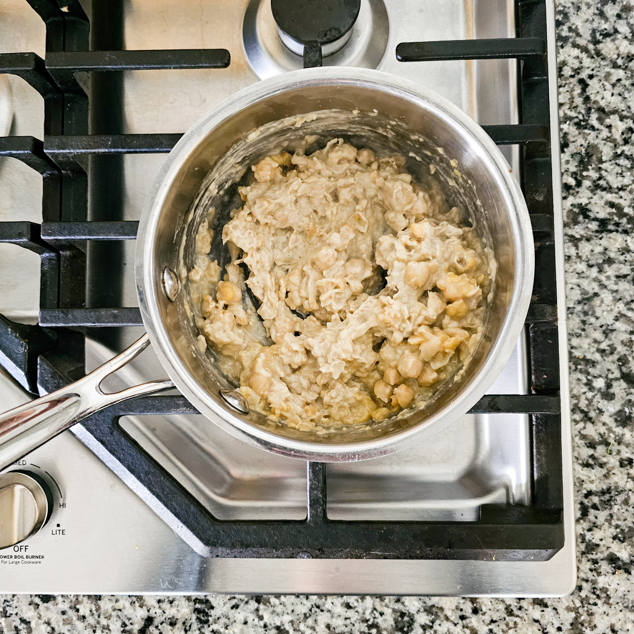 Ingredients to make the chickpea mixture of gluten-free vegan creamy musabaha stirred together in a stainless-steel saucepan on a gas cooktop.
